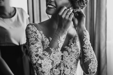 stylish happy bride putting on earrings and smiling, rustic wedding morning preparation in home. bridal getting ready. emotional moment. space for text. black white photo
