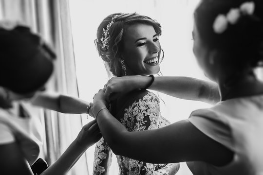 stylish happy bride dressing in dress, smiling to bridesmaids, at window, rustic wedding morning preparation. bridal getting ready. emotional moment. space for text. black white photo