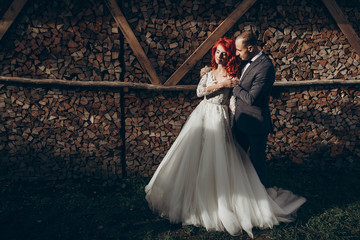 rustic wedding couple embracing in sunlight at background of wooden firewood. barn wedding concept, space for text. happy stylish bride and groom at wood wall, romantic sensual moment