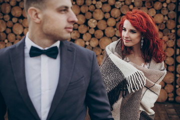 rustic wedding couple embracing posing at background of wooden firewood. barn wedding concept, space for text. happy stylish bride in blanket and groom at wood wall, romantic sensual moment