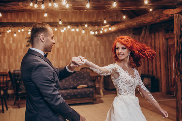 stylish groom and happy bride dancing under retro bulbs lights in wooden barn. rustic wedding concept, space for text. newlyweds couple embracing, sensual romantic moment