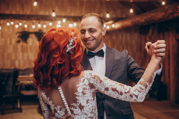 stylish groom and happy bride dancing under retro bulbs lights in wooden barn. rustic wedding concept, space for text. newlyweds couple embracing, sensual romantic moment