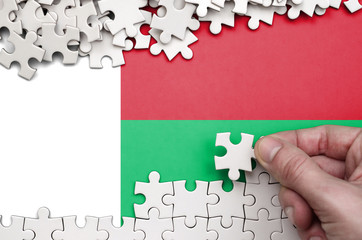 Madagascar flag  is depicted on a table on which the human hand folds a puzzle of white color