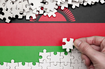 Malawi flag  is depicted on a table on which the human hand folds a puzzle of white color