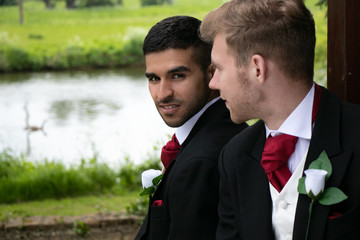 Gay couple of grooms pose for photographs by a lake on their wedding day