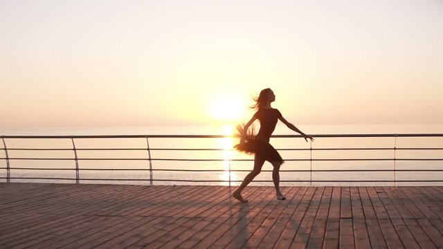 Young woman in black tutu doing ballet at the seafront. Bosk. Sunlight. Attractive ballerina practices in jumping. Side view. Slow motion