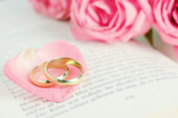 Golden wedding rings on pink rose petal with pink rose flower bouquet on open book with vintage tone