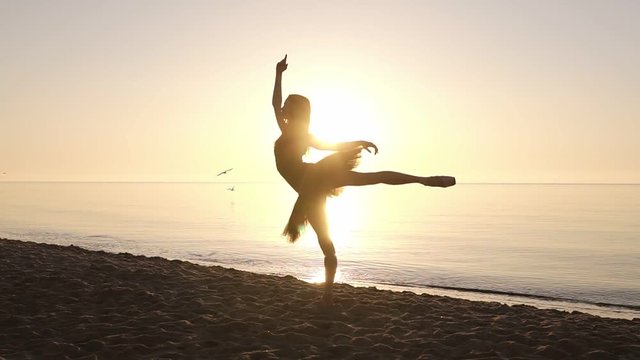 Silhouette of an elegant ballerina in a tutu in the rays of the morning sun. Doing exercises, practicing. Seashore. Slow motion