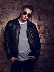 Male thinking model posing in leather jacket and trendy eyeglasses on brick wall street background....