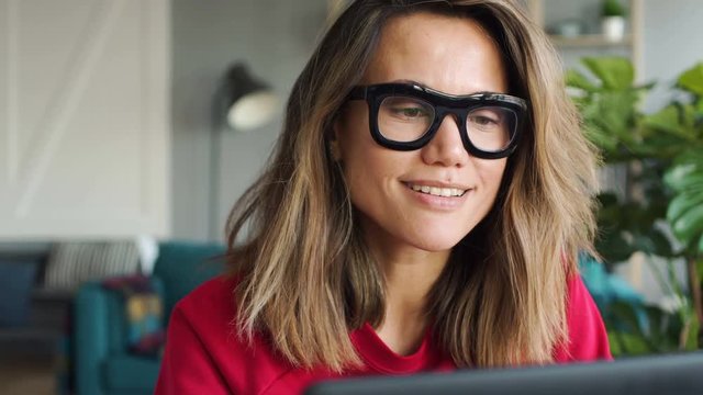 Close-up of attractive stylish caucasian woman in big glasses in her twenties concentrates on her studies and work on laptop at the table in the grey room in scandinavian style with golden bookcase