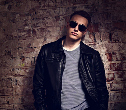 Male model posing in leather jacket and trendy eyeglasses on brick wall street background. fashion toned contrast portrait with empty copy space