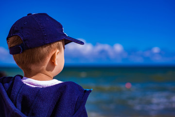 The boy looks at the sea. A small child in a hat is on the beach in the summer and looks at the waves of the sea.