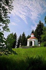 Vertical image of Saint John of Nepomuk rotunda of in pseudo-romanesque style standing on a green grass hill with white facade, red roof, cross, surrounded with coniferous trees, sunny day, blue sky