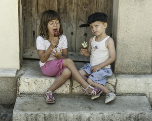 Portrait of two untidy scruffy dirty young caucasian french children blissfully eating ice-cream in front of rustic doorway in summer