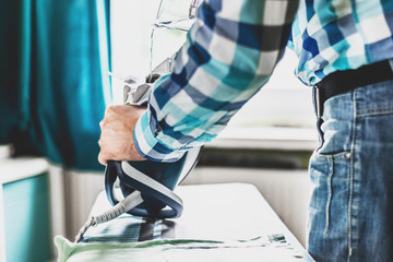 Man at home ironing clothes. Father performs home duties, irons the child's clothes. Man in role of a woman at home. Man ironing shirt on ironing board with steaming blue iron.