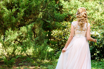 Fototapeta na wymiar Beautiful bride in wedding dress with bridal bouquet in the park outdoors, back view. Blond girl with curly hair styling and jewelry. Wedding hairstyle for long hair with stylish hair accessory