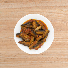 Serving of sprats in a tomato sauce on white plate atop a wood table.