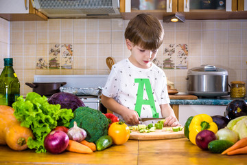 The young boy in cooking standing in the kitchen near table with vegetables
