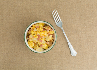 Top view of a bowl of scrambled eggs with potatoes cheese and ham plus a fork to the side on a brown tablecloth.