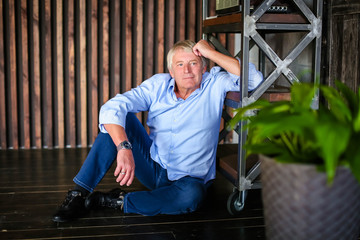 An elderly, joyful, gray-haired man in blue shirt, sits on floor, near rack in room