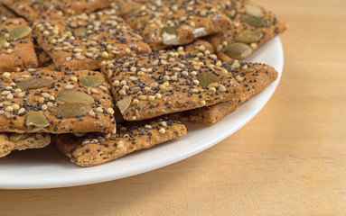 Side close view of whole wheat seeded crackers on a white plate atop a wood table.