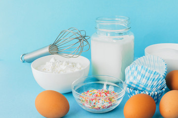 Eggs of chicken, flour in a bowl, sugar, milk in a glass jar whisk ingredient for cooking a bake on a blue background with copy space