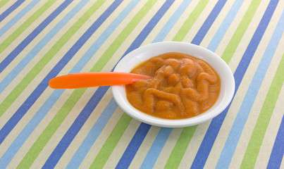 Bowl of organic pumpkin and carrot baby food with a spoon in the food on a striped tablecloth.