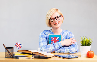 Smart female student, blonde woman wears eyeglasses, holding English language book on table,...