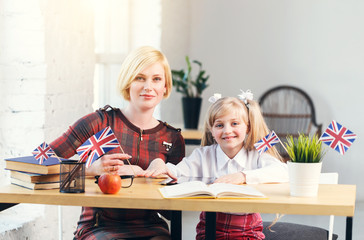 Female educator of English and smart girl smiling, working table with books, british flag and apple, kids education language concept