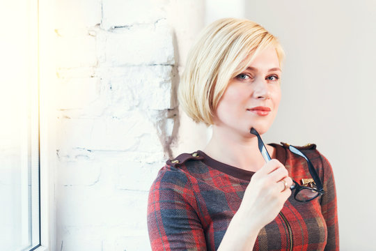 Pretty intelligent blonde woman wears eyeglasses and red dress reading book near wide window in laght room
