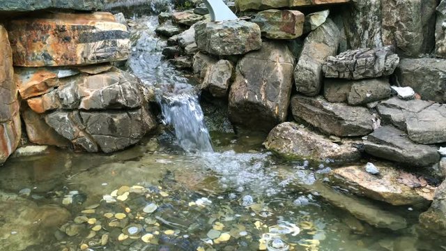 Current clear water stream between stones, or mini-waterfall, which falls into a pond with small coins on the bottom. On a sunny day.