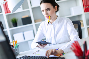 A young girl in the office holds a pen in her mouth and works with a calculator, documents and a...
