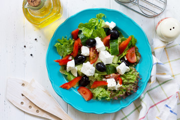 Greek salad with fresh vegetables, feta cheese and black olives in a blue plate on white wooden table. Top view flat lay background.