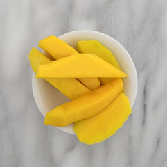 Slices of fresh mango in a white bowl atop a gray marble counter top.