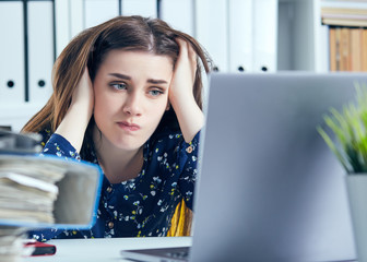 Young tired and exhausted woman looks at the camera leaning against mountain of documents propping up her head with her hands.