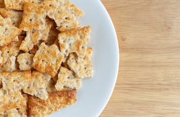 Top close view of broken flatbread salted multigrain crackers on a white plate atop a wood table.