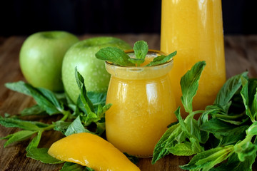 Yellow smoothie in glass jars surrounded by fruits and mint against wooden background