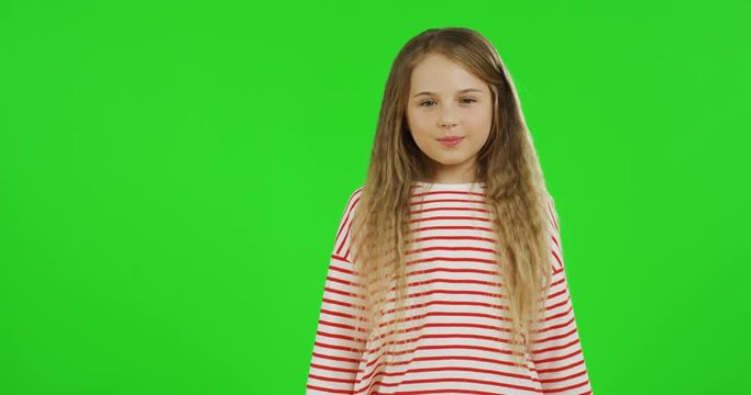 Funny small schoolgirl with long hair standing on the chroma key background, showing her tangue to the camera and crossing her hands. Green screen.