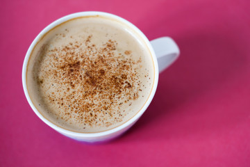 Morning Ñappuccino. Cup of coffee with milk foam and cinnamon or cacao powder on bright pink background.