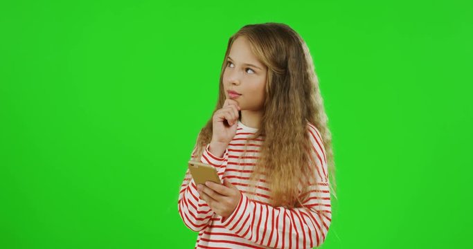 Adorable small teen girl with long fair hair standing on the chroma key background, thinking and scrolling on the smartphone screen. Green screen