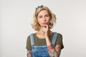 Realy? Studio shot of cute blonde female keep her finger under chin, weras denim overalls, thinking...