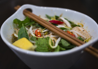 Beautiful and appetizing photo of a traditional vietnamese chicken noodle soup, also know as Pho Ga. In a white bowl with wooden background and luxurious wooden chopsticks.