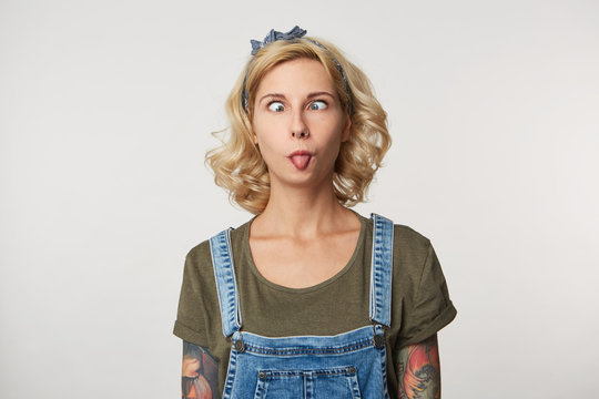 Indoor shot of young female has fun, makes grimace and shows tongue, weras casual denim overalls and t shirt. Portrait of funny young woman being crazy