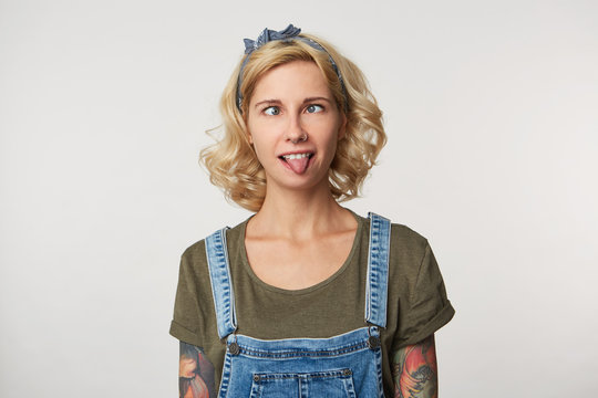 Indoor shot of young female has fun, makes grimace and shows tongue, weras casual denim overalls and t shirt. Portrait of funny young woman being crazy