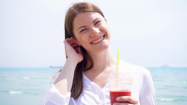 Smiling vegan woman in white shirt holding cup with strawberry smoothie against sea in slow motion. Fit vegetarian female enjoying healthy lifestyle outdoors