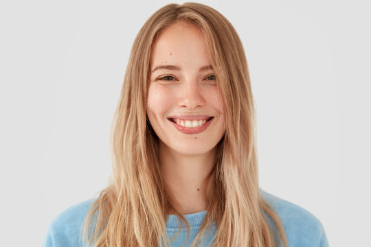 Close up portrait of good looking European female hears something pleasant, has broad smile and white teeth, has light long hair, poses against white background. People, lifestyle, facial expressions
