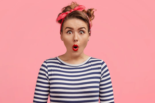 Omg, I can`t believe my eyes! Beautiful pinup girl in striped sailor turtleneck sweater, hears unbelievable news, says wow with amazement, stands against pink background. Emotive woman being excited