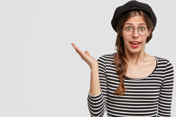 Beautiful French woman in stylish beret, striped turtleneck sweater, indicates with palm on left copy space, poses against white background. Attractive amazed female shows something wonderful