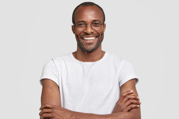 Portrait of unshaven pleased dark skinned male has warm broad smile, keeps hands crossed, rejoices...