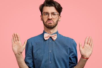 It`s not my business! Serious honest male worker keeps palms raised in front, shows that he is not about of this, wears elegant jean shirt with pink bowtie and spectacles. People and body language